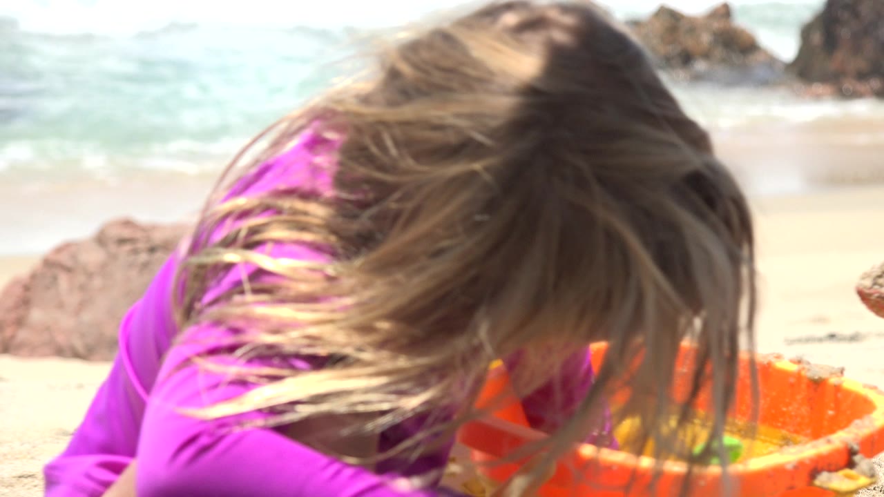 The child leans farther toward the orange bucket, her hair covering her face as sunlight hits the sand and calm water in Cabo San Lucas.