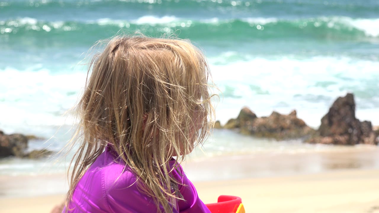 Blond child in a purple top beside ocean surf and beach rocks.