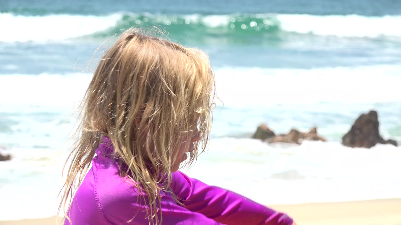 Blonde child in a purple beach top fills the left foreground on a Cabo San Lucas beach, with white surf and dark offshore rocks behind.
