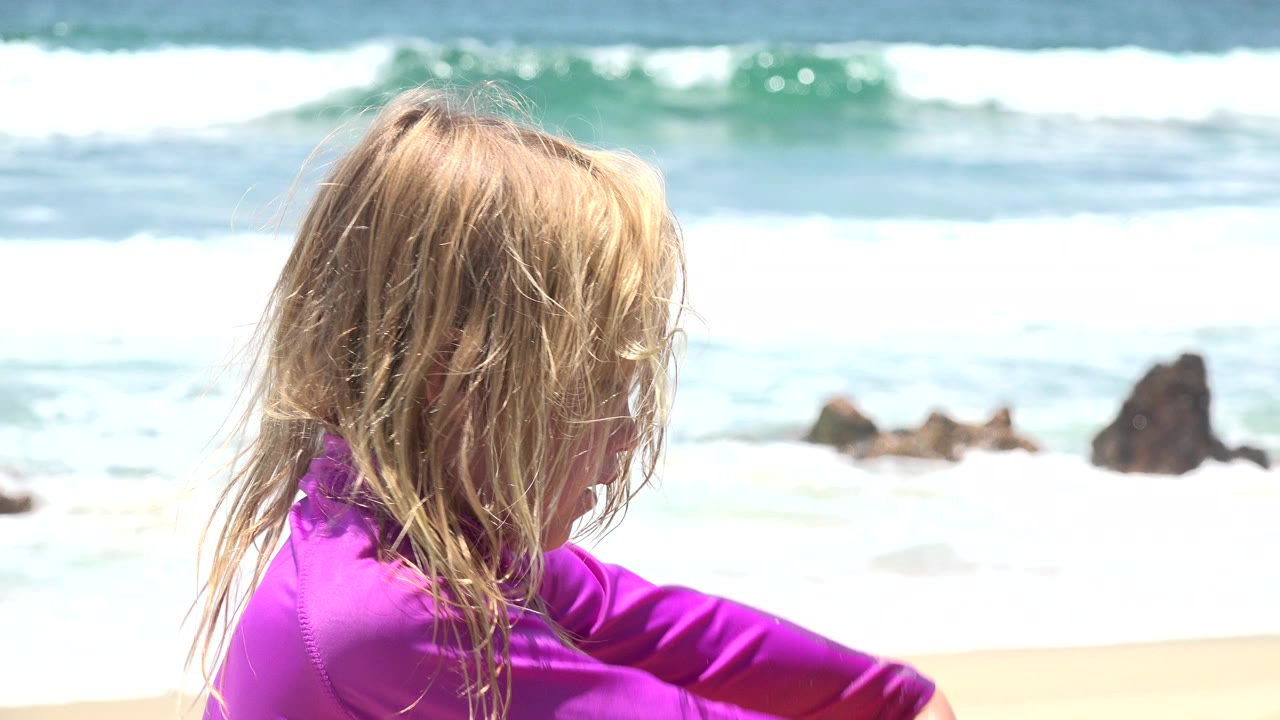The child remains in close side view, looking downward near the sand while bright surf rolls across the Cabo San Lucas shoreline in back.