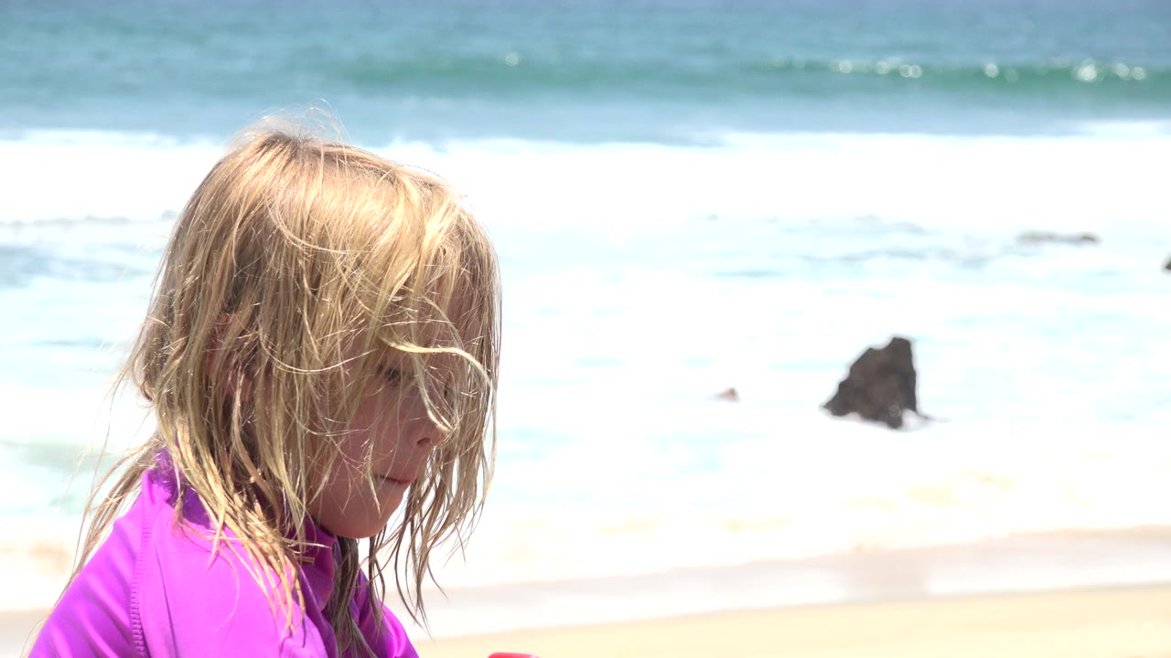 The child remains seated in the foreground on the Cabo San Lucas beach, looking down as windblown hair covers much of the face and surf rolls behind.