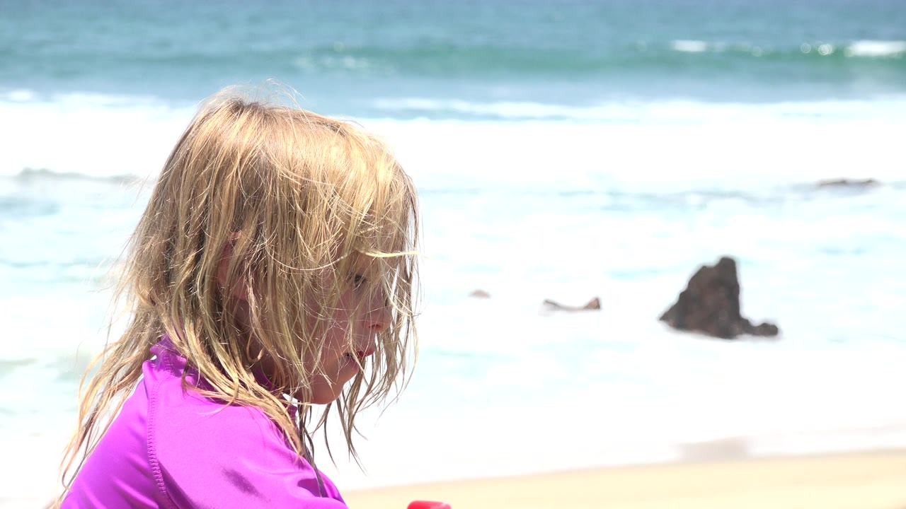 The child is shown in partial side profile on the right side of the Cabo San Lucas beach frame, with pale sand, foamy surf, and dark rocks in the background.