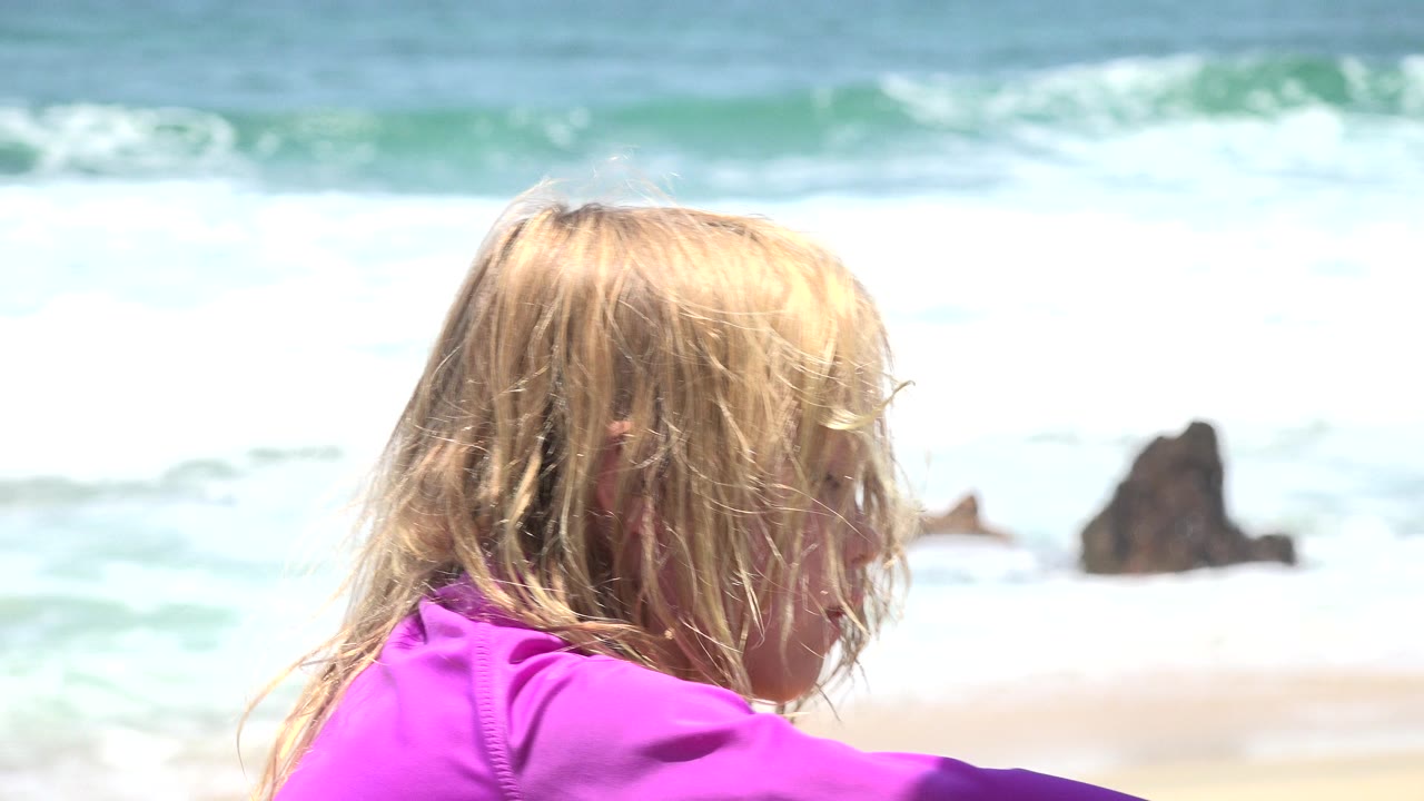 Blond child in a purple top near ocean surf.