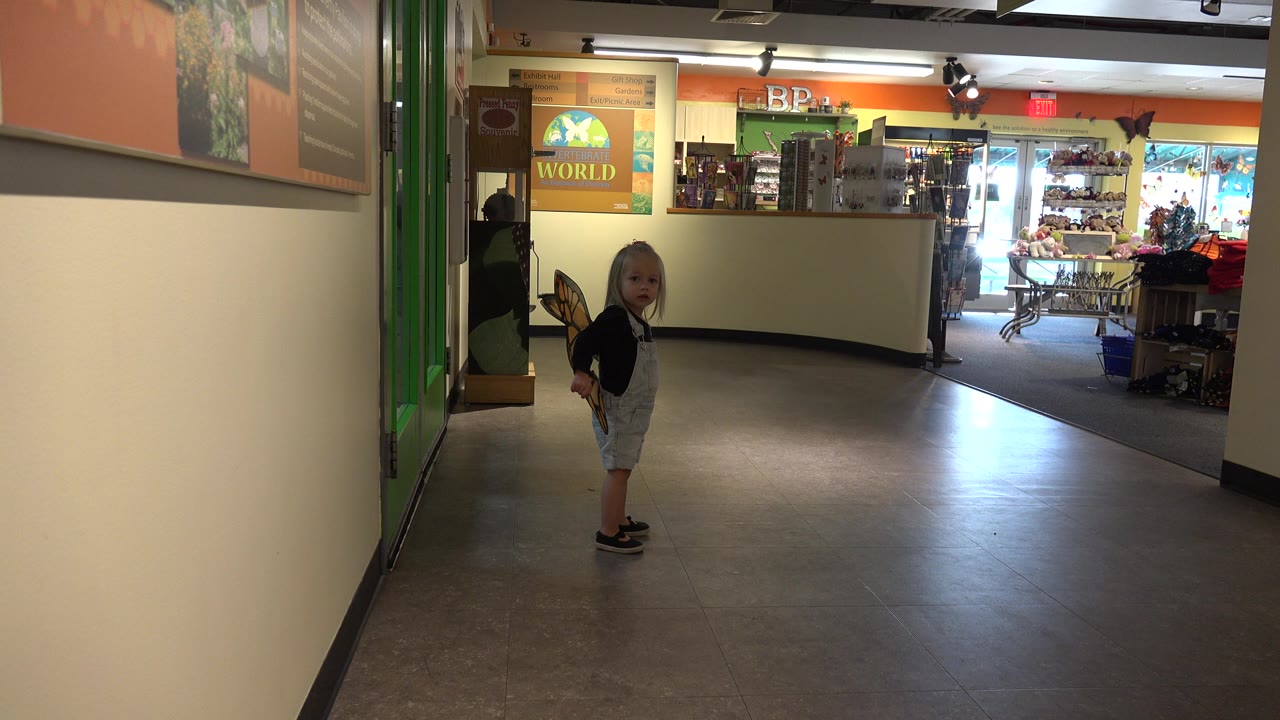 The child in butterfly wings remains in the midground of a Broomfield indoor corridor, facing the camera with wall signs and a bright background area visible.