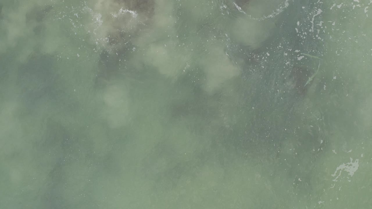 Overhead view of blue-green ocean water in Baja California Sur, Mexico, with faint white foam near the top edge, cloudy submerged patches, and no visible people.