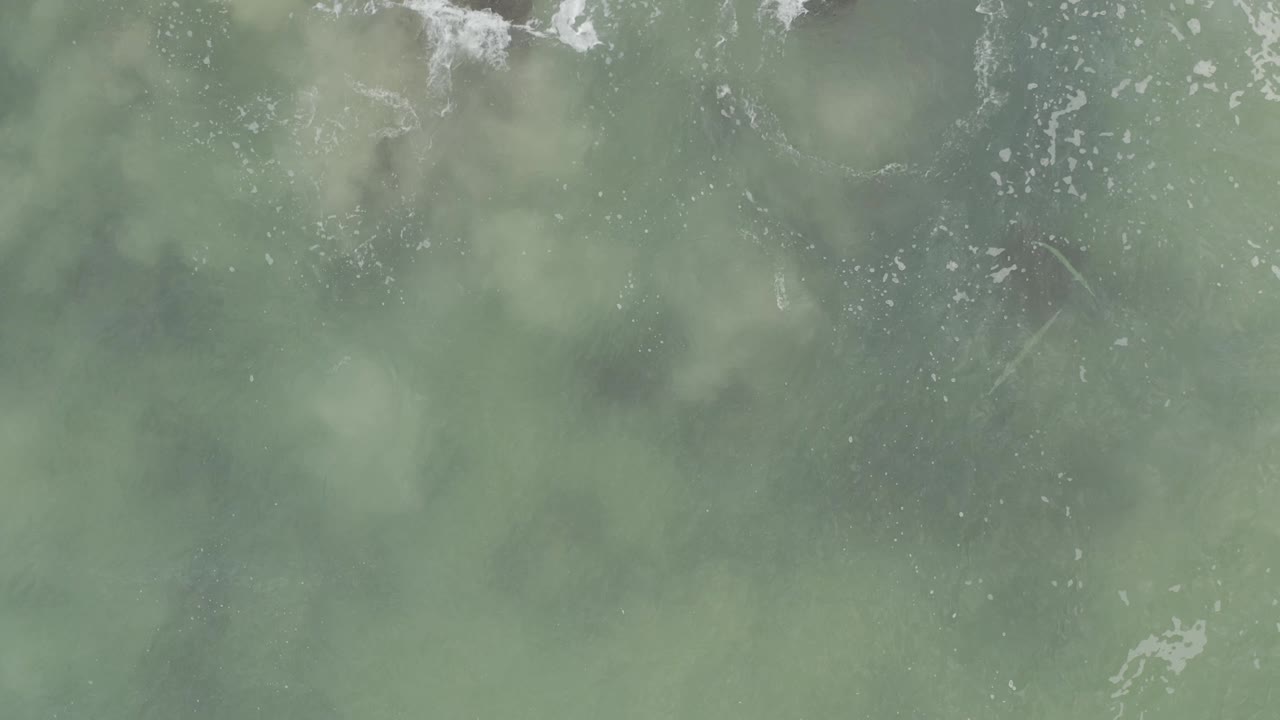 Blue-green ocean surface in Baja California Sur viewed from above, with pale foam streaks crossing the upper area and diffuse submerged forms below, with no visible people.