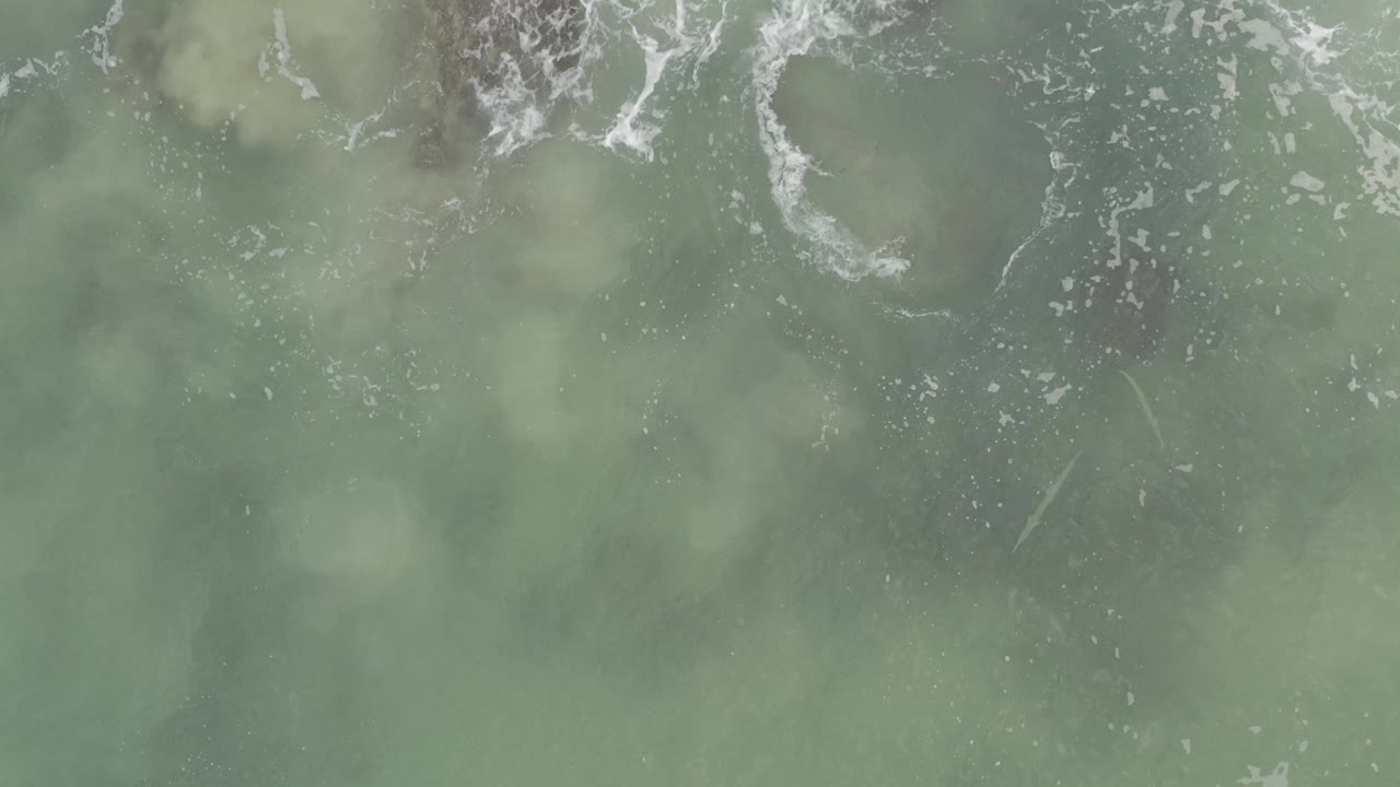 Overhead Baja California Sur ocean frame with brighter white foam breaking across blue-green water and cloudy green submerged patches, with no visible people.