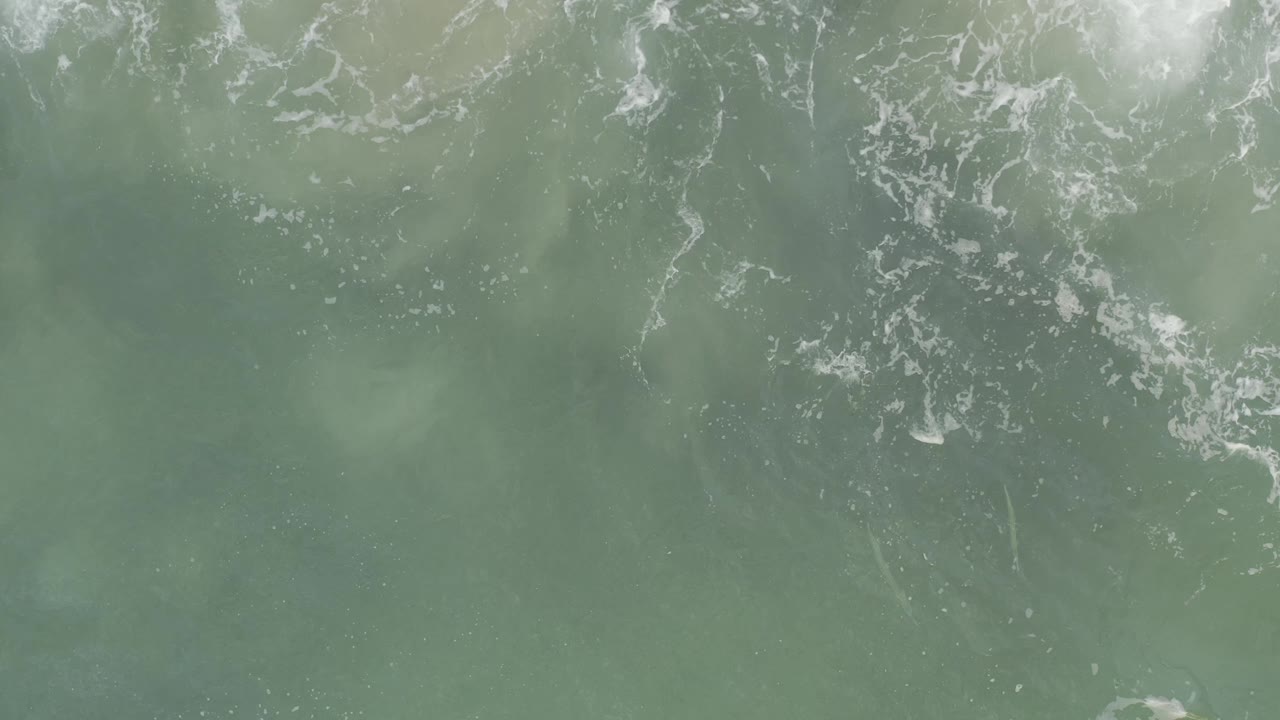 Top-down Baja California Sur ocean surface with pale blue-green water, small white foam clusters along the top and right side, and no visible people.