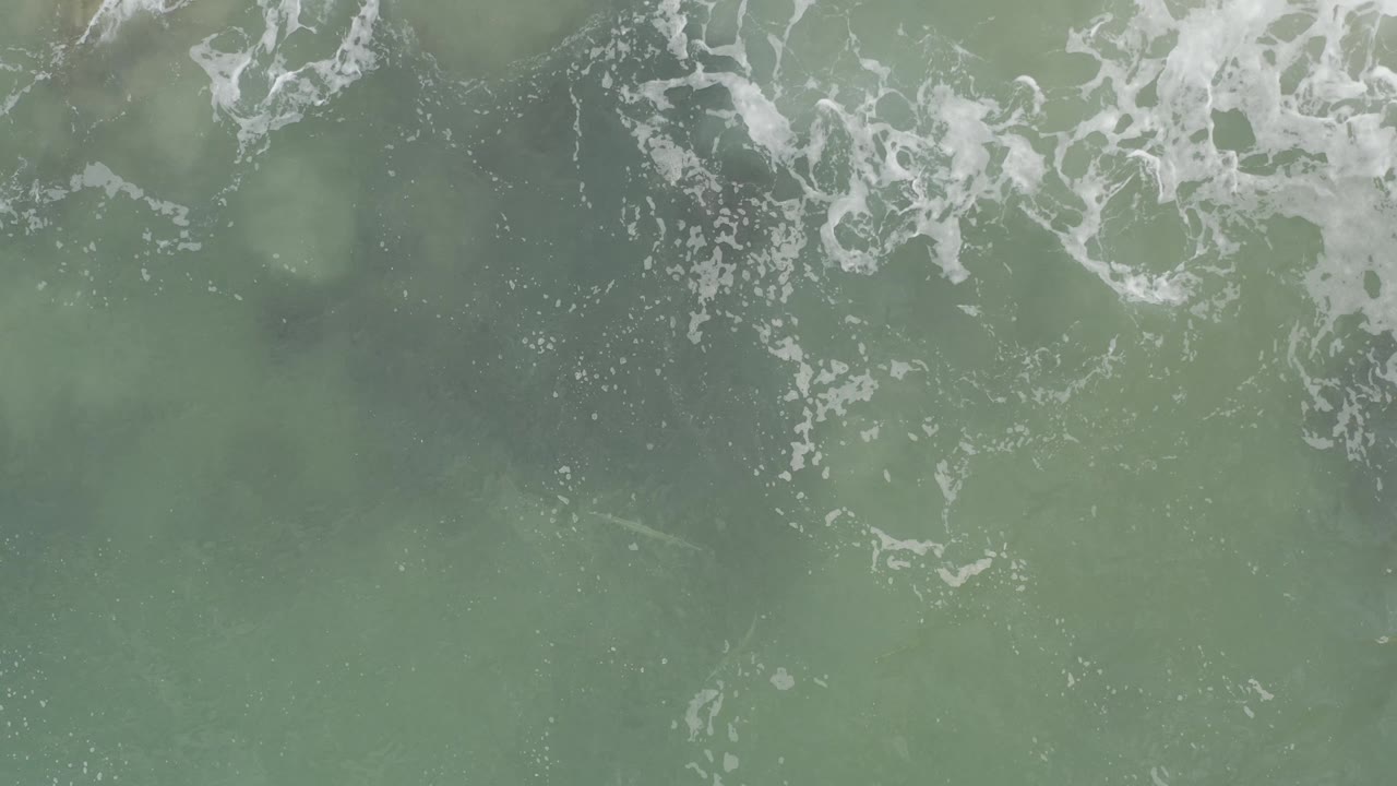 Top-down view of muted green ocean water in Baja California Sur, Mexico, where white foam breaks across the upper edge and no visible people appear.