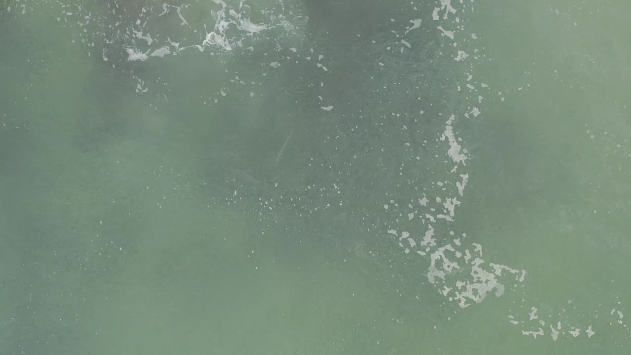 Blue-green shallow water fills the frame in Baja California Sur, with scattered foam lines and a darker underwater patch near the center, no visible people.