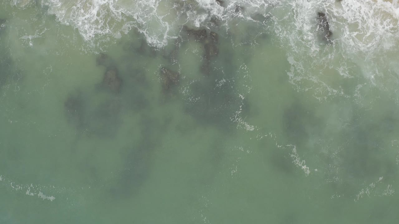 Overhead aerial view in Baja California Sur, Mexico, with white surf crossing dark rocks under blue-green water and no visible people.