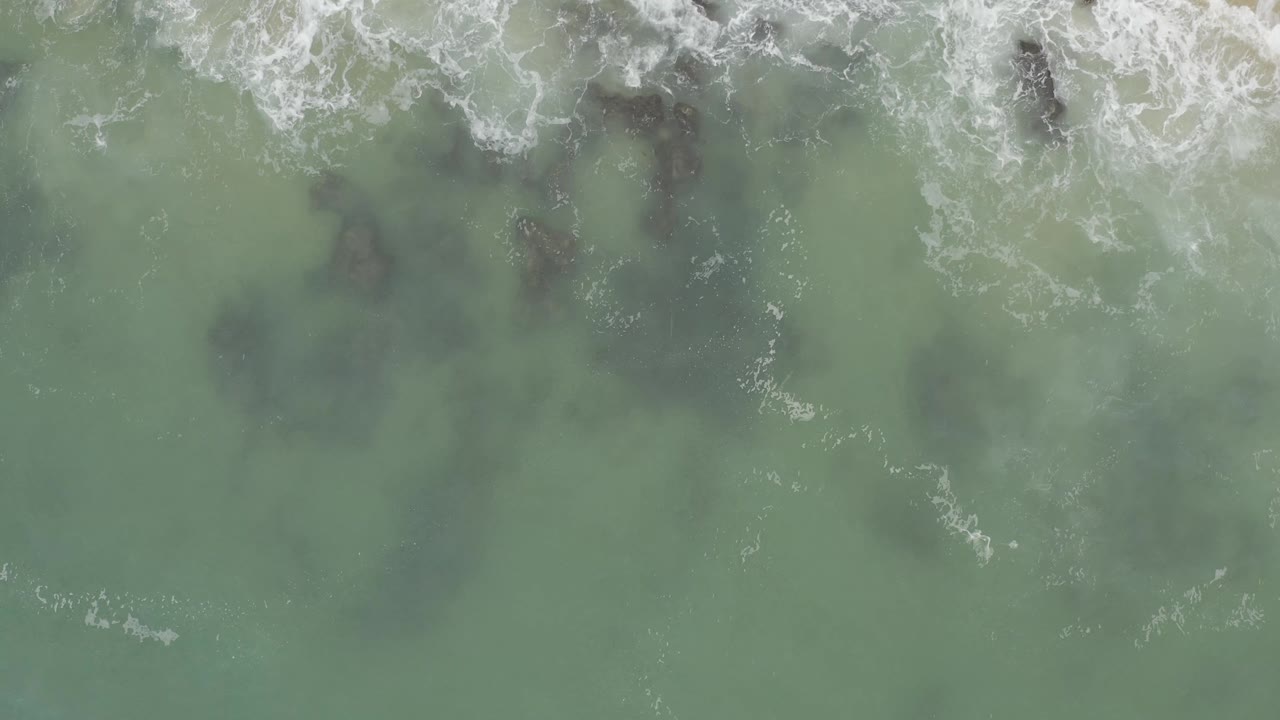 Aerial frame over Baja California Sur surf, with white foam thickening along the upper edge and dark submerged rocks below, no visible people.