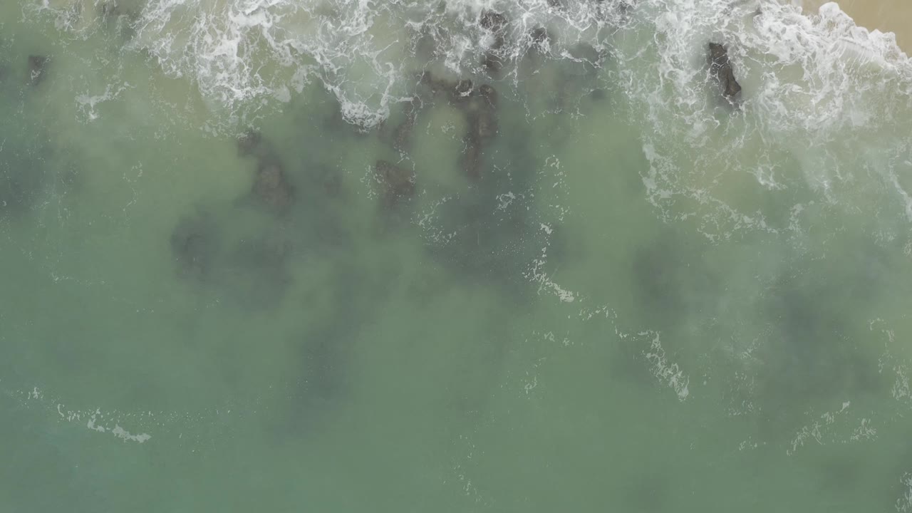 Top-down Baja California Sur coastline view with pale green water, scattered dark rocks beneath the surface, and foamy surf at the top, no visible people.