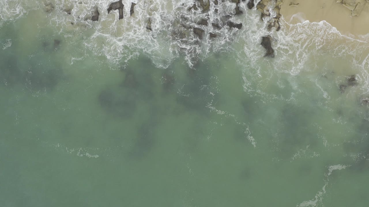 Overhead view in Baja California Sur, Mexico, with pale green water, white surf, dark rocks, tan sand along the top edge, and no visible people.