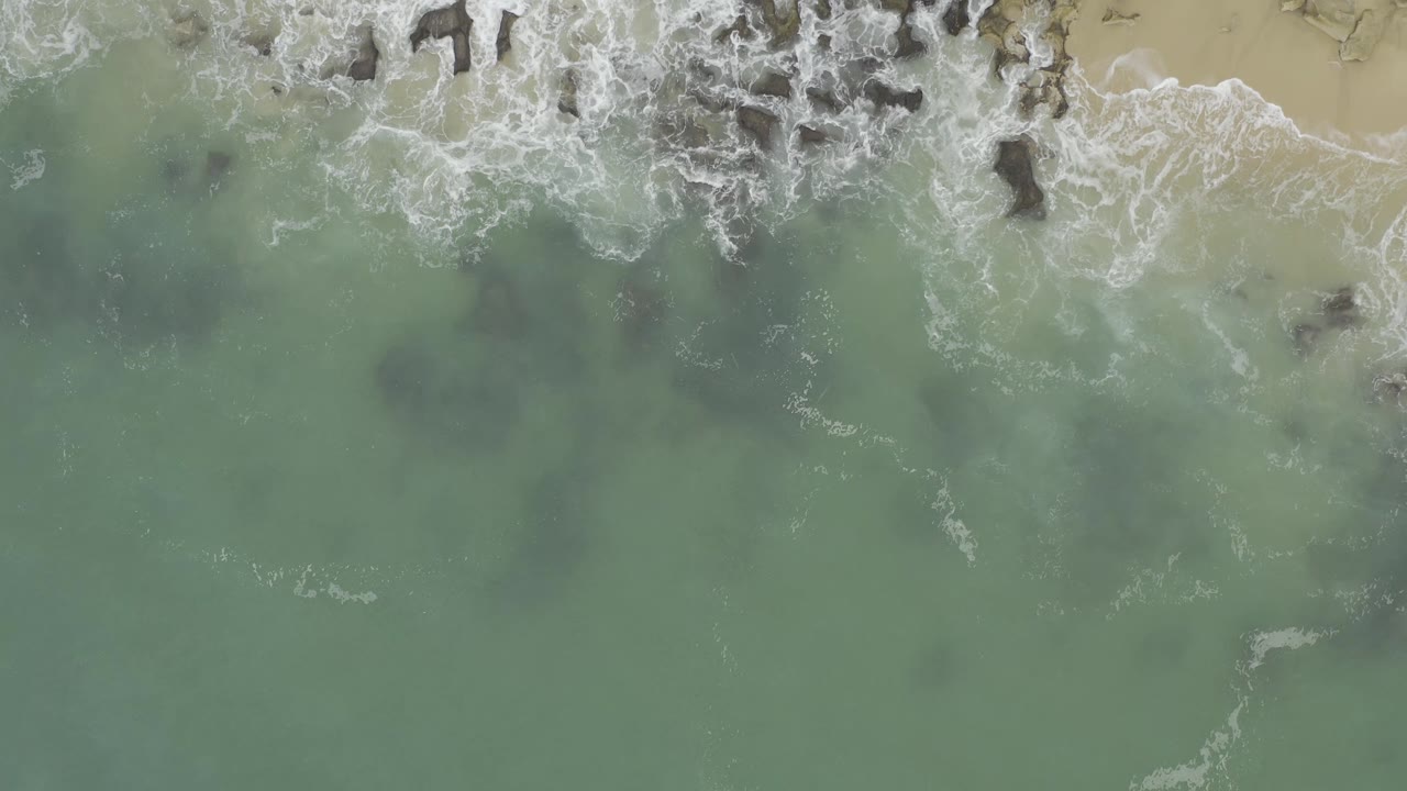 Aerial frame in Baja California Sur, Mexico, showing white foam spreading over dark nearshore rocks with green water below and beach at the top right, with no visible people.