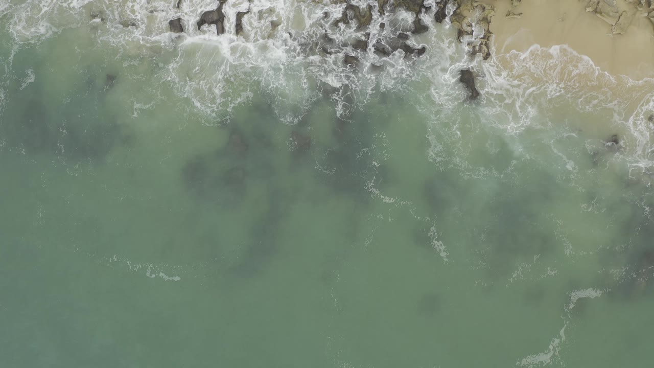 Top-down Baja California Sur coastline frame with green water occupying most of the image, white surf across the upper edge, scattered dark rocks, and no visible people.