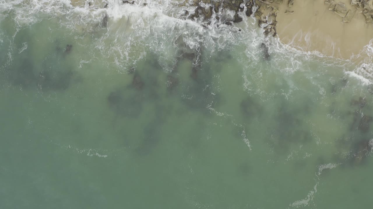 Aerial view in Baja California Sur, Mexico, with a larger white wave wash crossing shallow green water, dark rocks beneath the surface, sand at the upper right, and no visible people.