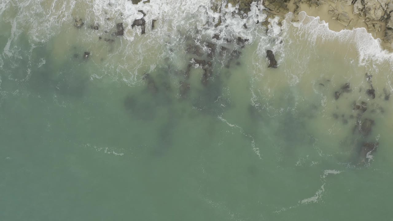 Aerial frame of the Baja California Sur coast with white foam spreading across shallow green water and scattered dark rocks beneath the surface, with no visible people.