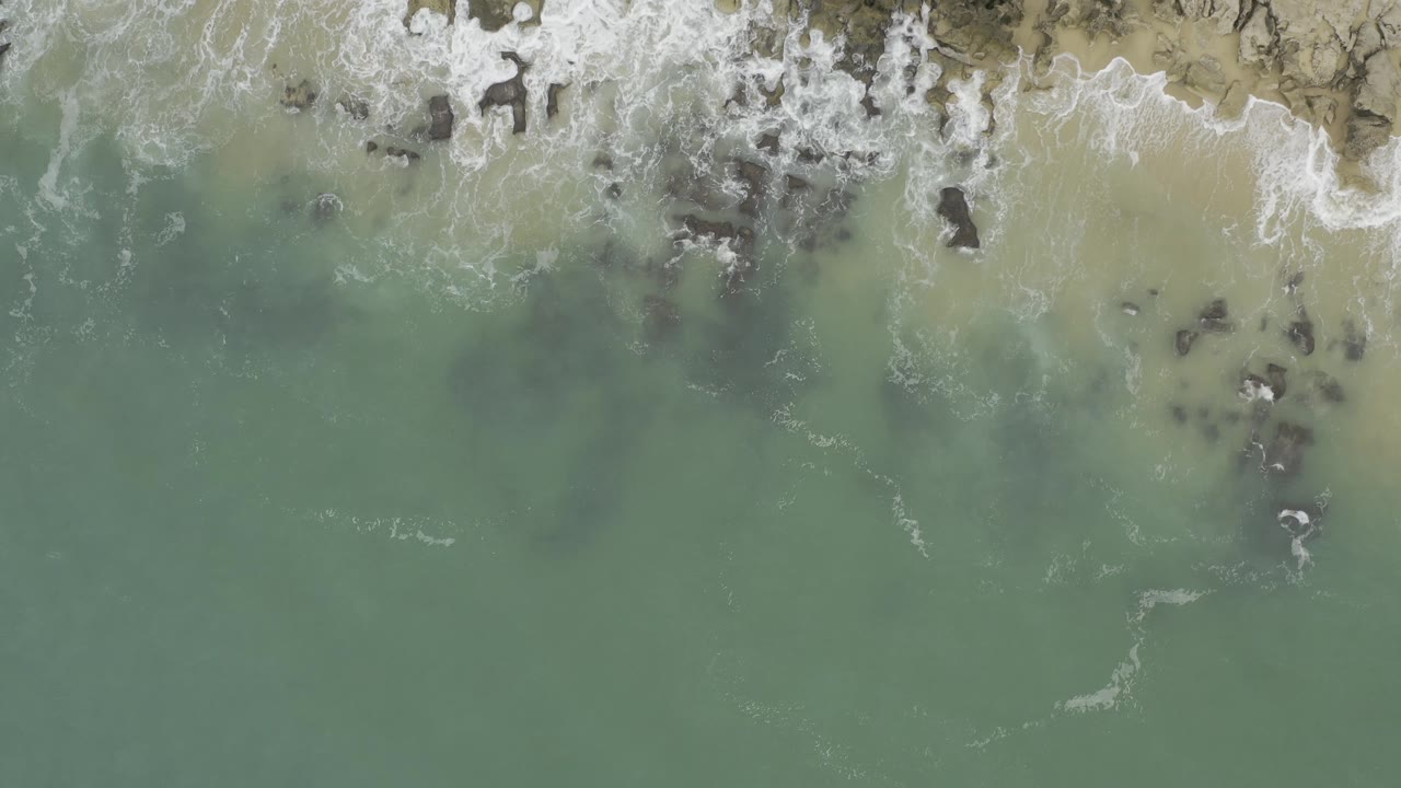 Top-down Baja California Sur shoreline view with surf curling across the top of frame, tan sand on the right, and muted green water over dark rock patches, with no visible people.
