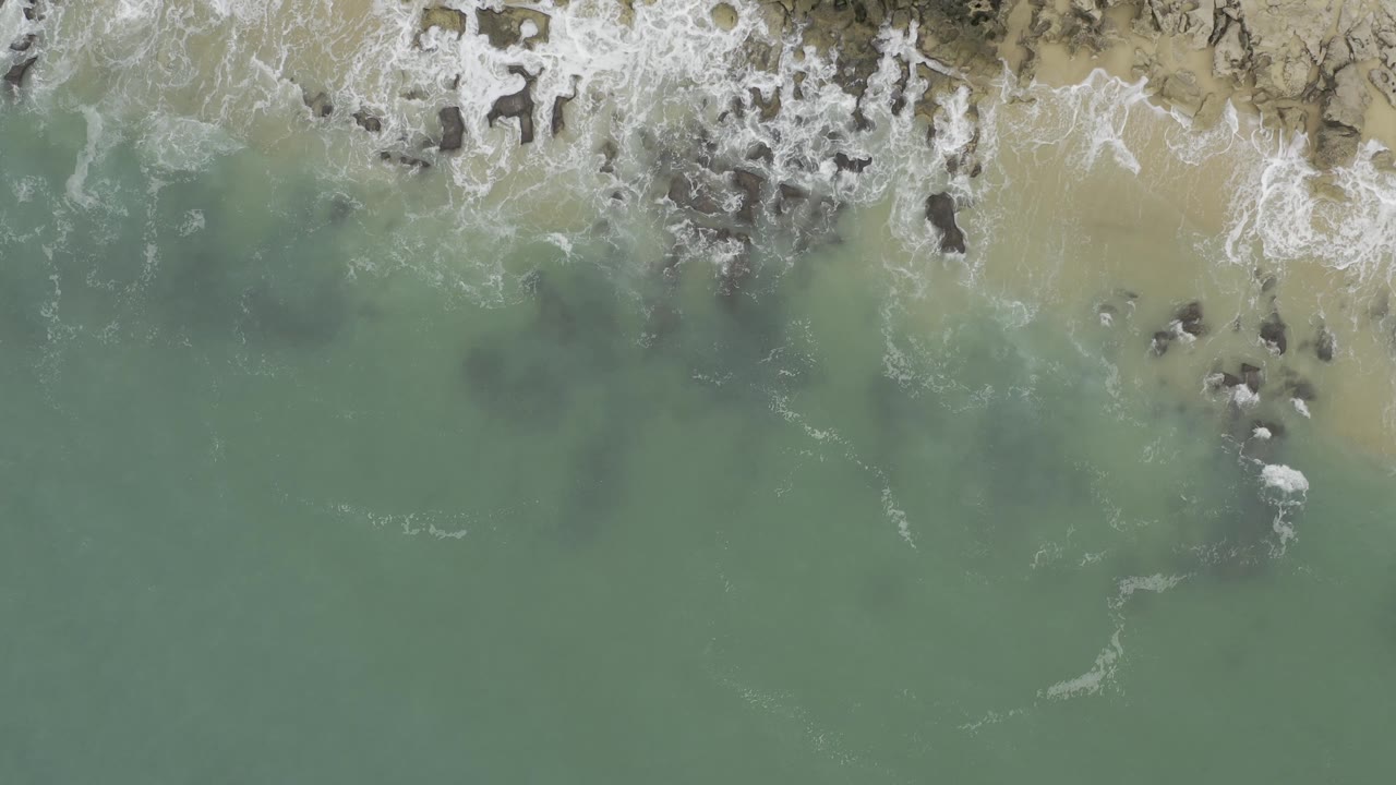 Straight-down aerial view of Baja California Sur surf breaking across pale sand and clustered dark rocks, with cloudy green water filling the lower frame and no visible people.