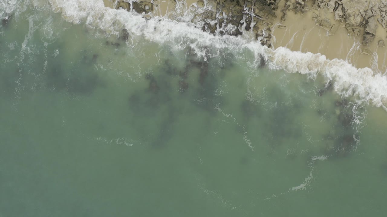 Overhead frame from Baja California Sur, Mexico showing green ocean water below white surf breaking across tan sand and dark rocks, with no visible people.