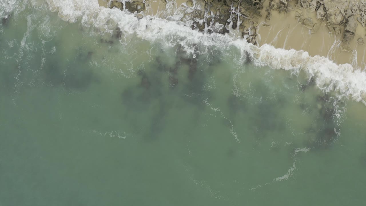 Aerial view in Baja California Sur, Mexico with a broad band of white foam crossing rocky tan shoreline above muted green water, with no visible people.