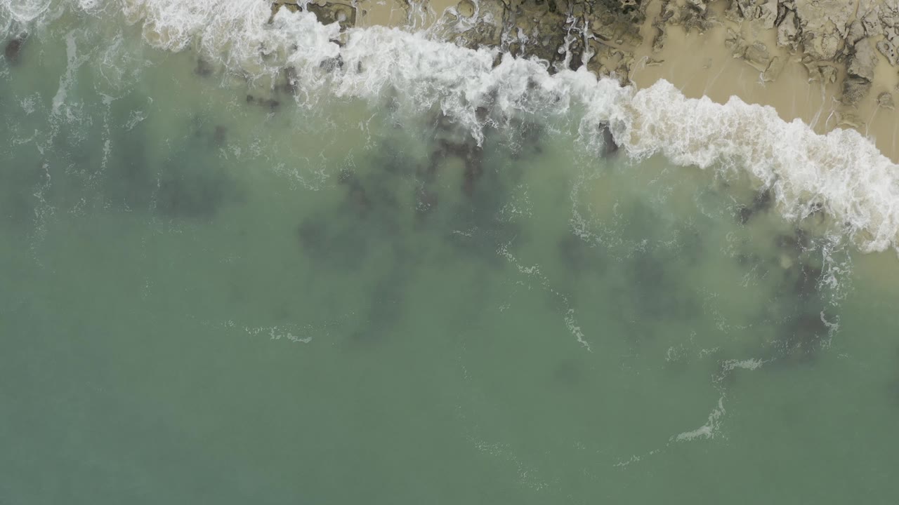 Top-down Baja California Sur shoreline view with green water in the lower frame and foamy white breakers along sand and exposed rocks near the top, with no visible people.
