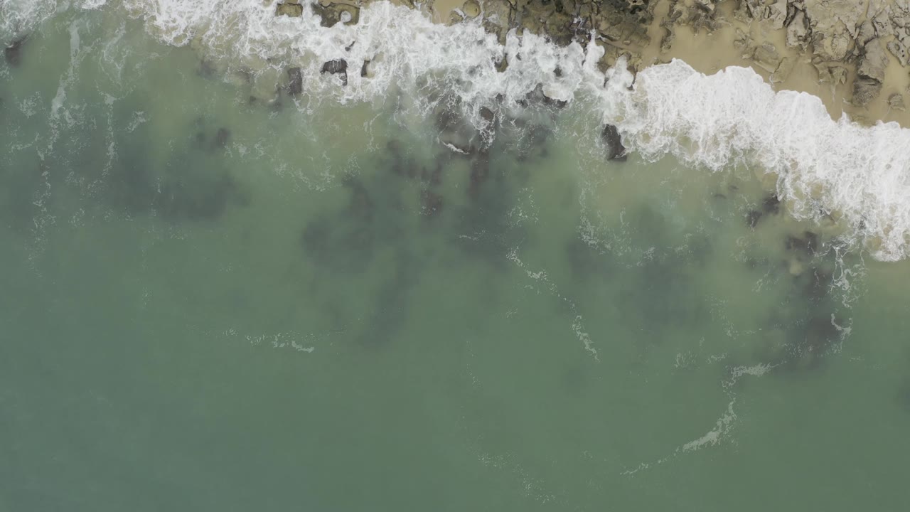 Overhead coastal frame in Baja California Sur, Mexico where white foam curls around dark rock shapes under shallow green water near a tan sandy edge, with no visible people.