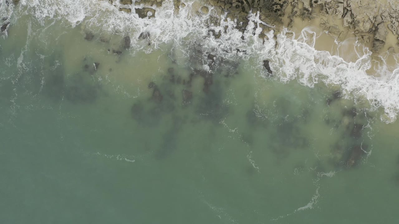 Top-down aerial frame in Baja California Sur, Mexico with a wider band of white foam along the top edge, dark underwater rocks near center, green water, and no visible people.