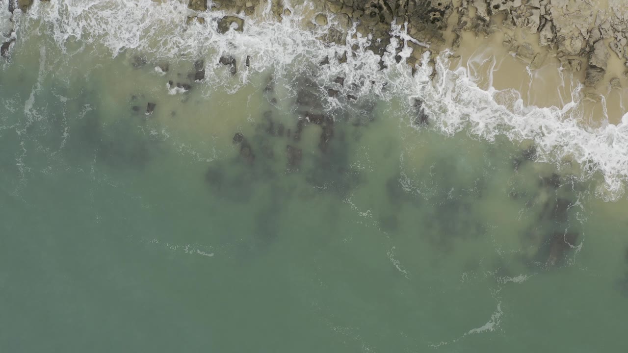Top-down aerial frame in Baja California Sur, Mexico showing clear green water below a foamy rocky beach edge, scattered dark rock forms, and no visible people.