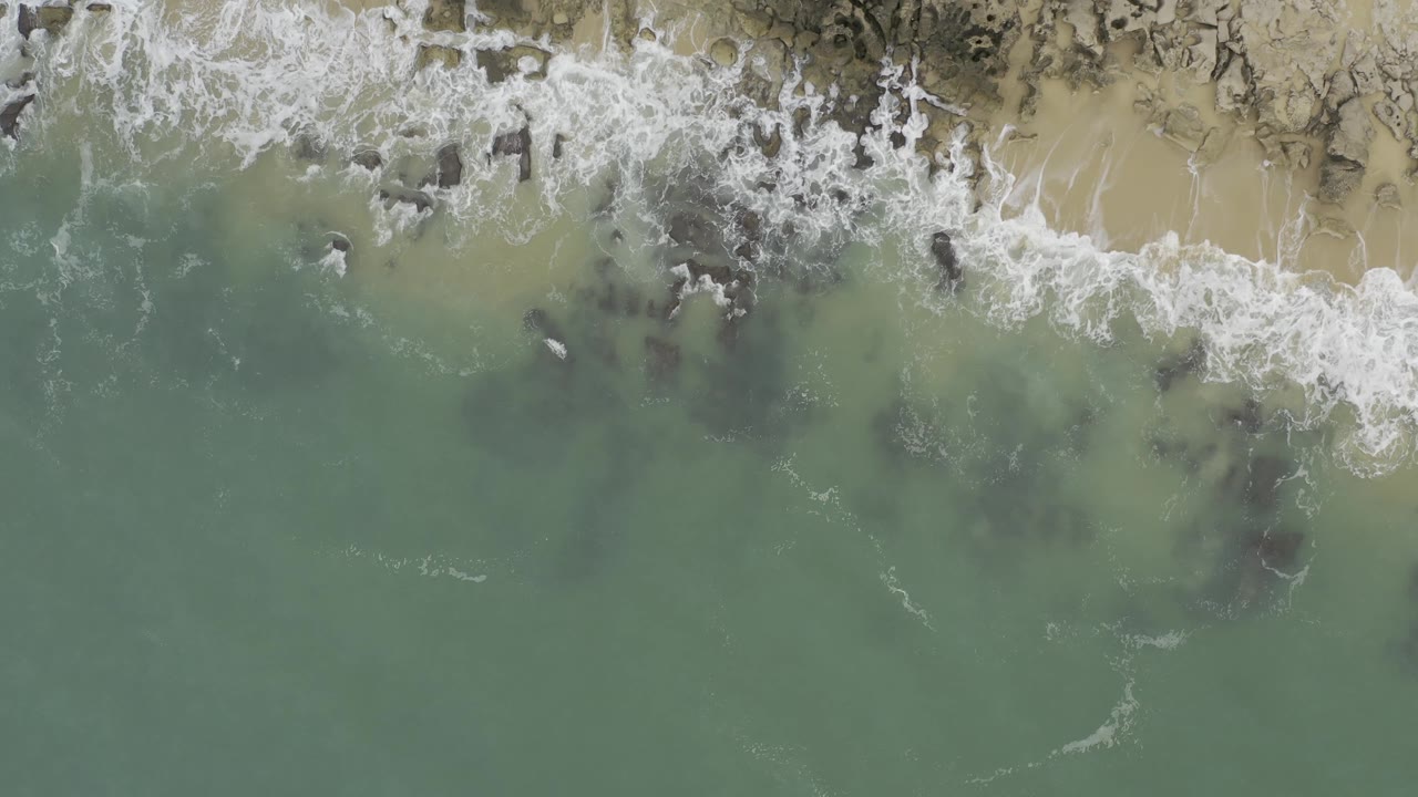 Top-down aerial frame in Baja California Sur, Mexico where surf washes over a cluster of rocks near pale sand, with green water below and no visible people.