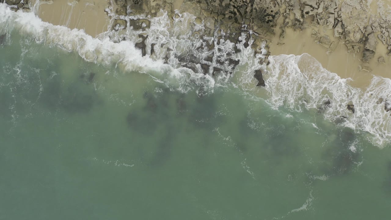 Overhead view of green water and white surf meeting pale sand and dark rocks in Baja California Sur, Mexico, with no visible people.