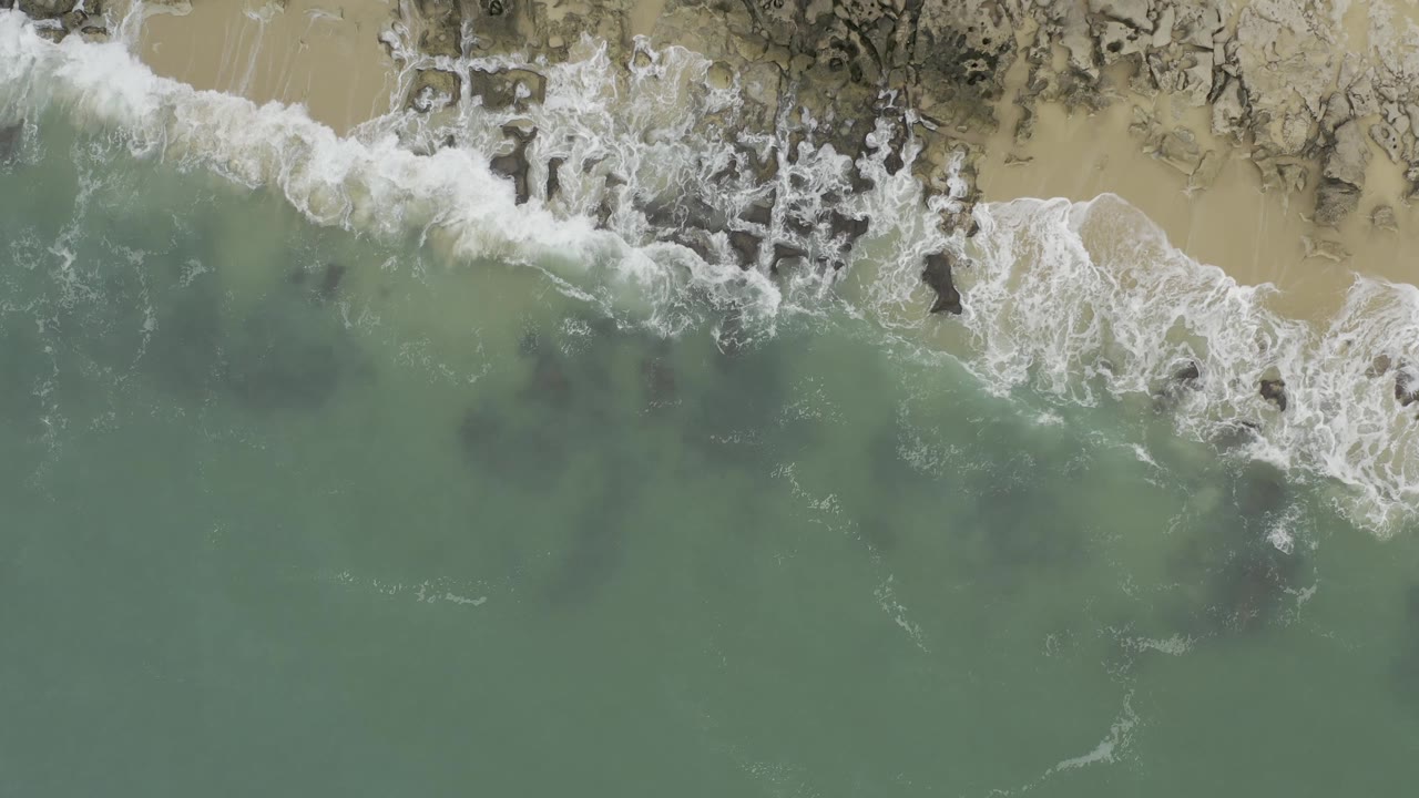 Green sea water sits below a band of white foam and pale beach in Baja California Sur, Mexico, with dark rock patches visible and no visible people.