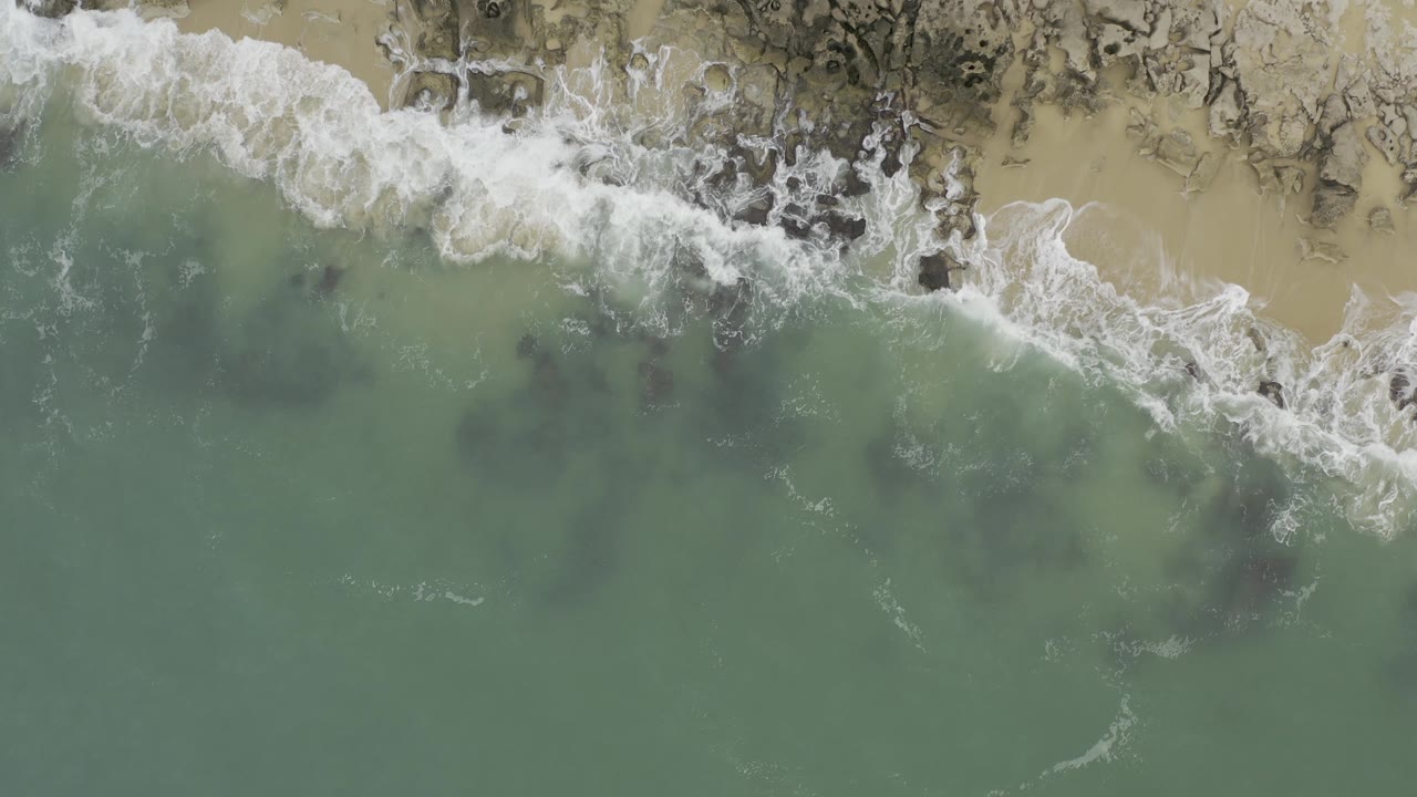 A diagonal strip of breaking white surf runs across sand and rock while green water fills the lower frame in Baja California Sur, Mexico, with no visible people.