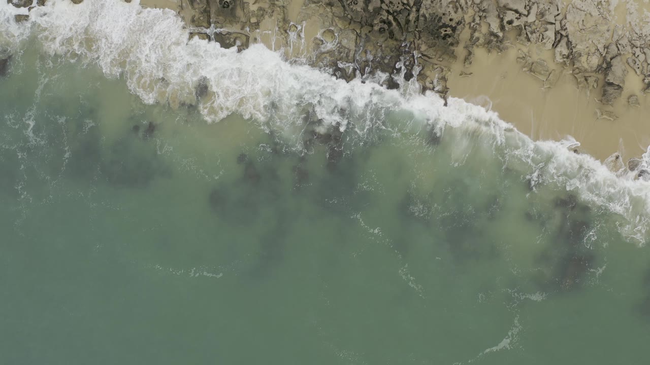 White foam pushes farther across the upper shoreline while muted green water and darker submerged patches remain below in Baja California Sur, Mexico, with no visible people.