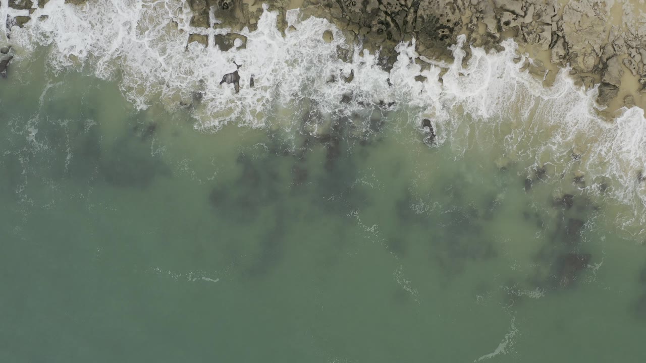 Top-down view of Baja California Sur shoreline with white surf breaking over dark rocks and green-blue water below, with no visible people.