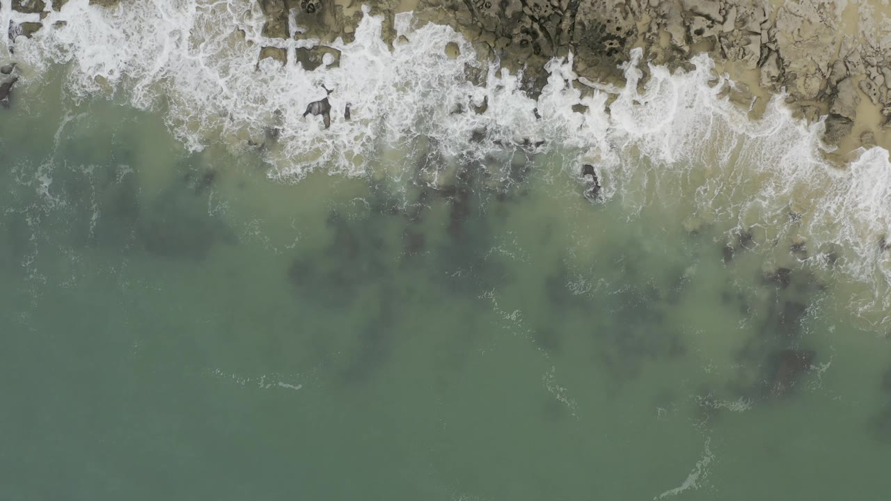 Aerial frame over Baja California Sur coast where foamy white waves spread along the rocky upper edge and darker submerged shapes sit in green water, with no visible people.