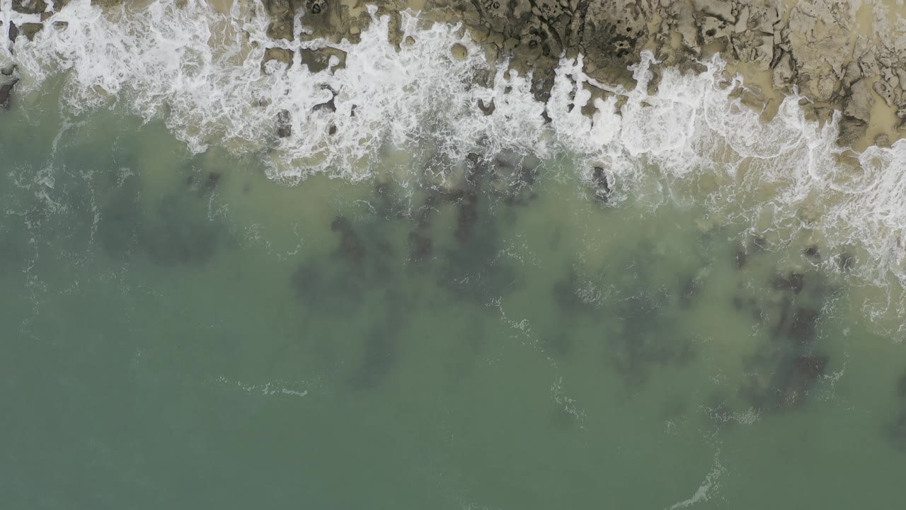 Top-down Baja California Sur shoreline view with a band of white surf at the top and broad green water below showing dark submerged rock patches, with no visible people.