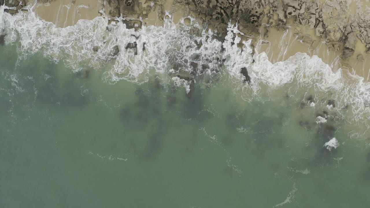 Overhead surf washing across sand and dark shoreline rocks.