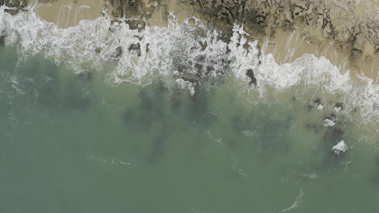 Aerial frame of pale surf foam spreading through blue-green shallow water toward tan beach in Baja California Sur, Mexico, with no visible people.