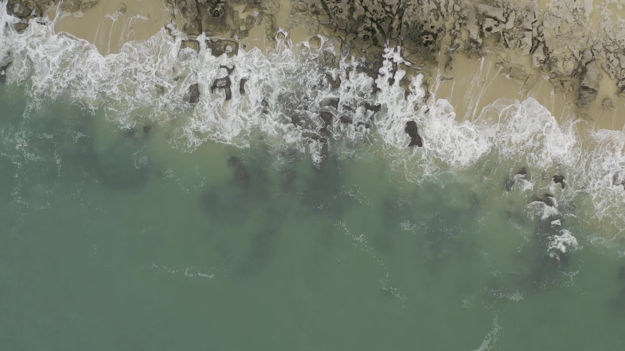 Top-down shoreline view where whitewater breaks across dark nearshore rocks and tan sand in Baja California Sur, Mexico, with no visible people.