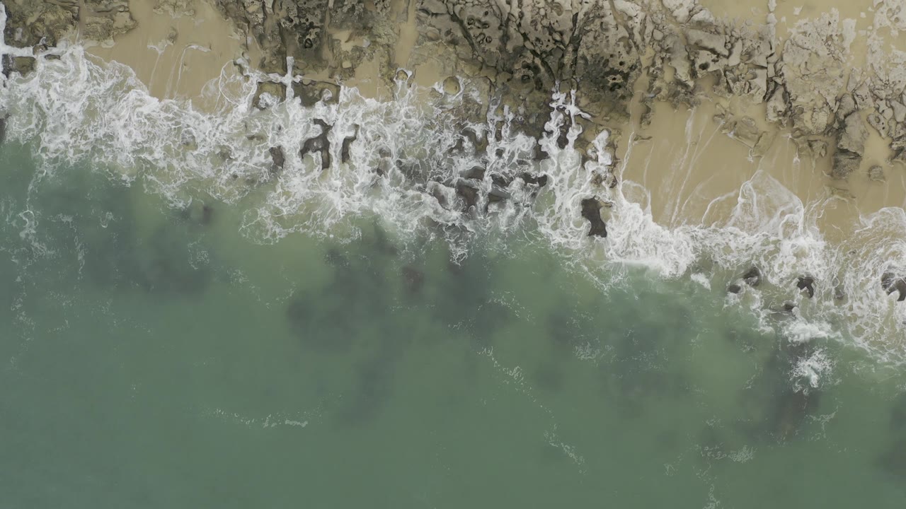Overhead coastal frame with green water below and white foam curling over dark rock patches along the sandy shore in Baja California Sur, Mexico, with no visible people.