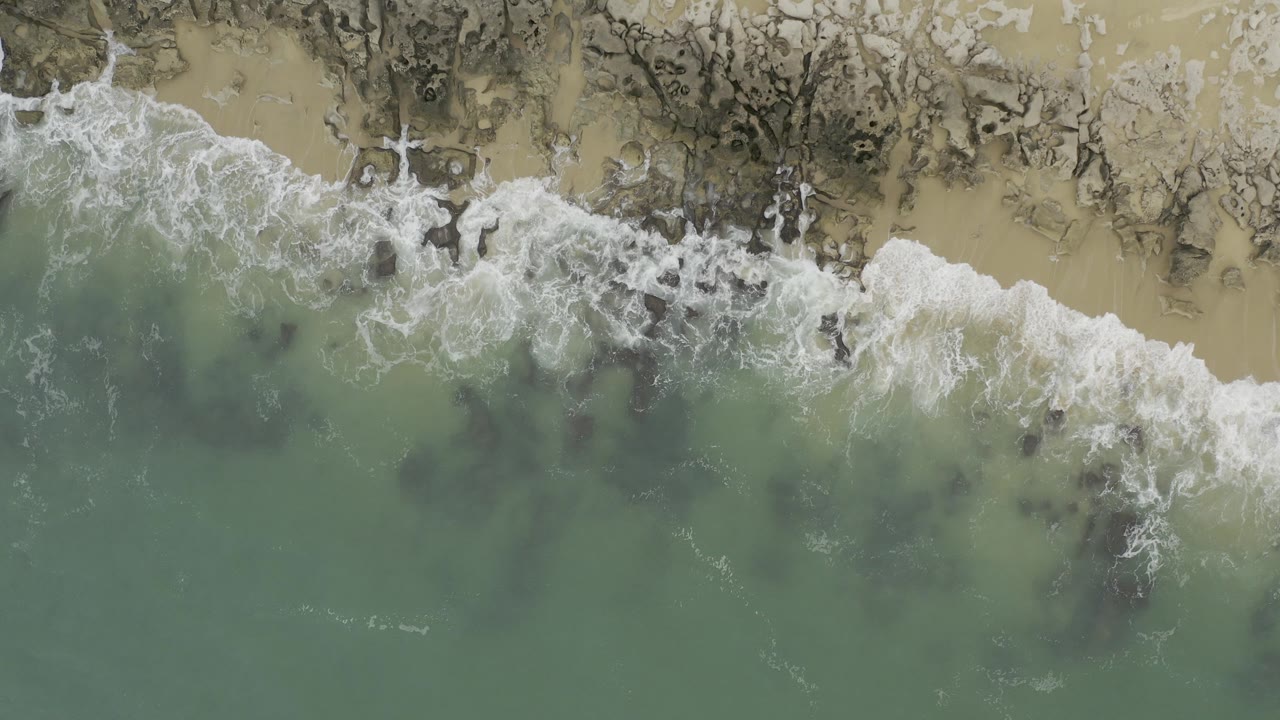 Overhead view of green water and white surf meeting a rocky sand shoreline in Baja California Sur, Mexico, with no visible people.