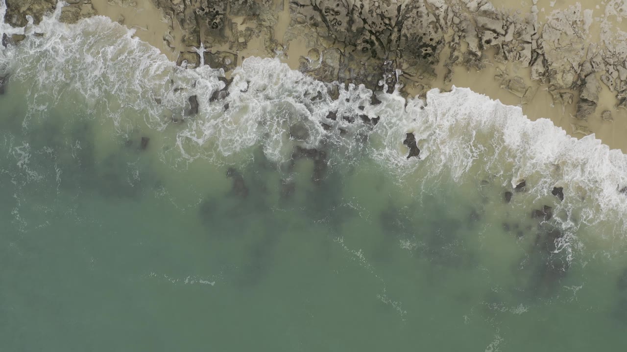 Top-down coastal view in Baja California Sur, Mexico, where curved white surf crosses dark rocks and a thin sand strip, with no visible people.