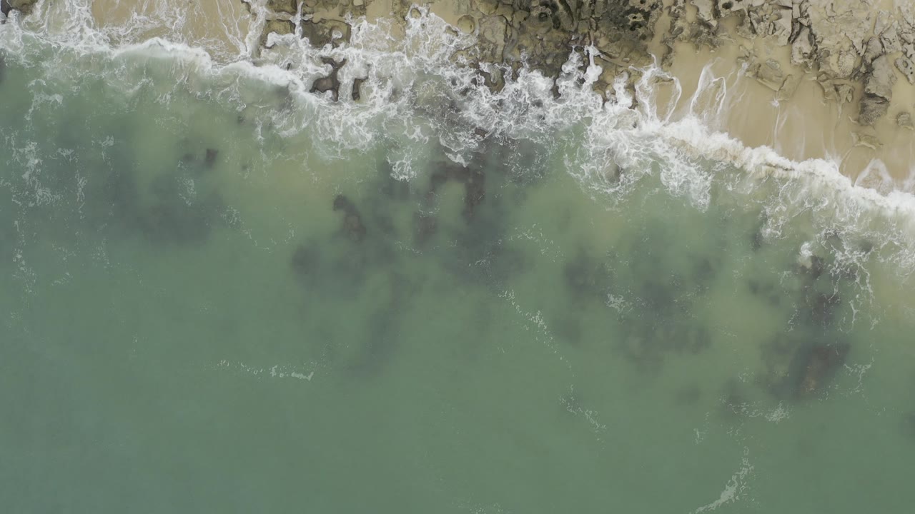 Overhead view of Baja California Sur surf washing onto pale sand and dark coastal rocks, with green-blue water below and no visible people.