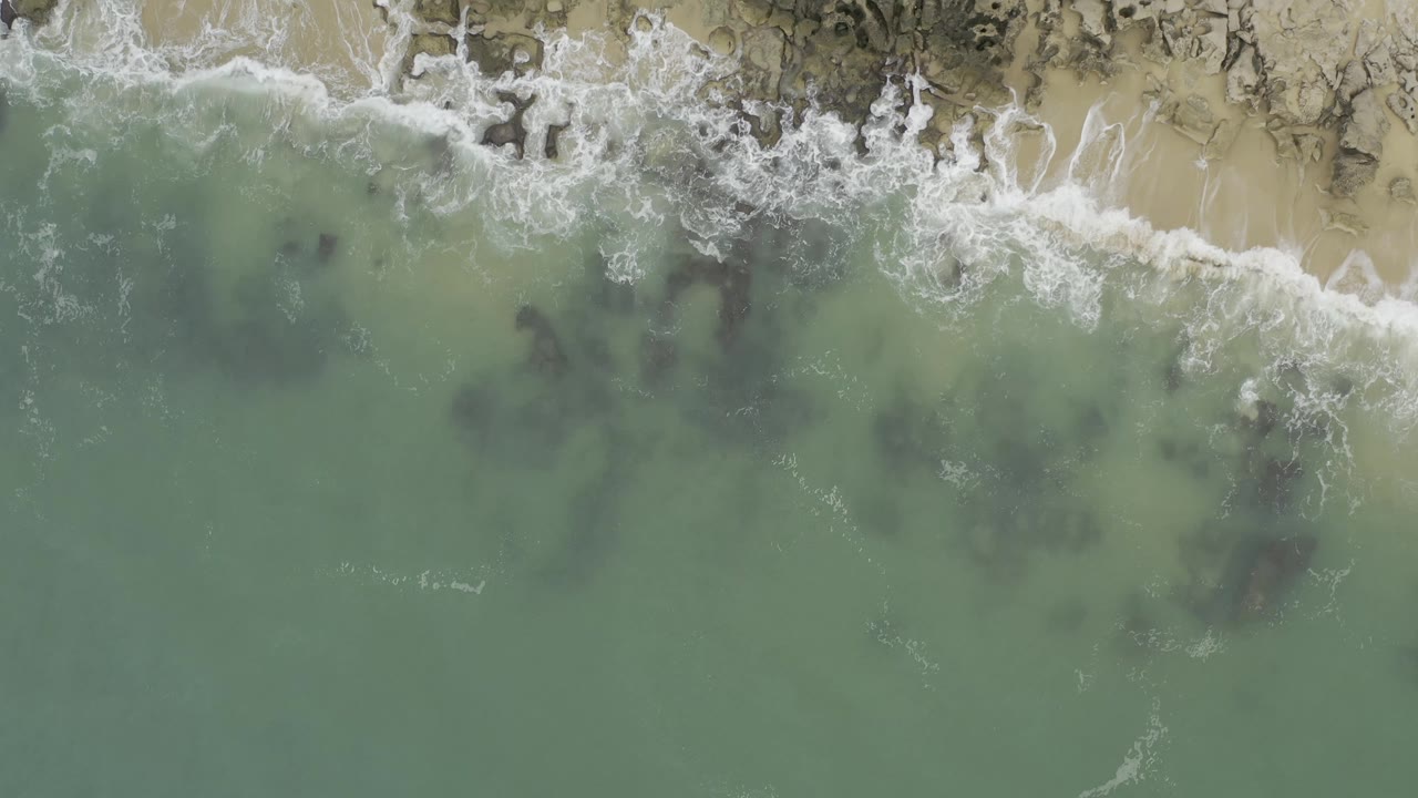 Aerial Baja California Sur shoreline frame with turquoise water, scattered dark rock patches, white foam at the beach edge, and no visible people.