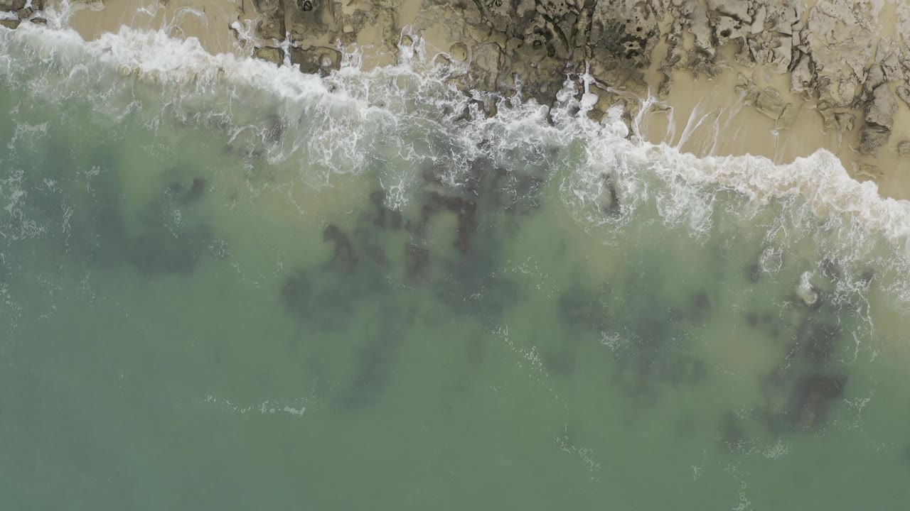 Top-down view of Baja California Sur waves breaking over a narrow sandy edge and dark rocks, with open green water filling the lower frame and no visible people.