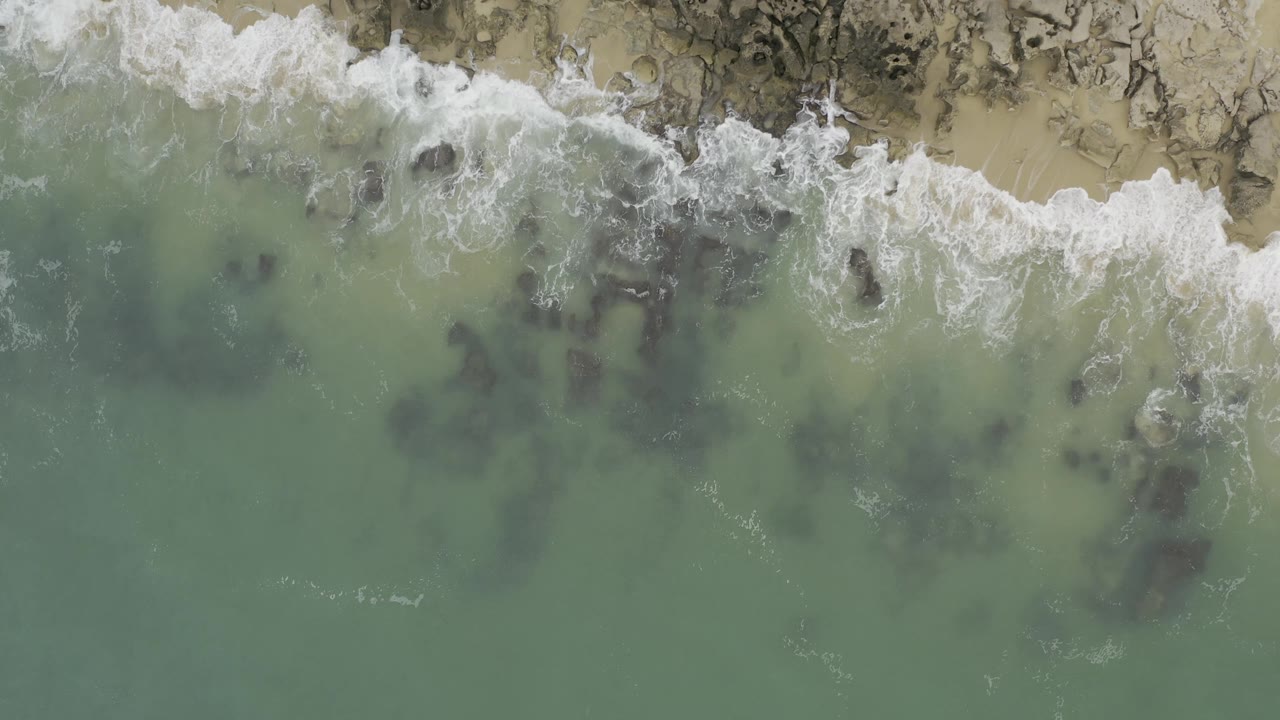 High aerial Baja California Sur coast view with white foam spreading over shallow rocks, pale beach at the top edge, green water below, and no visible people.