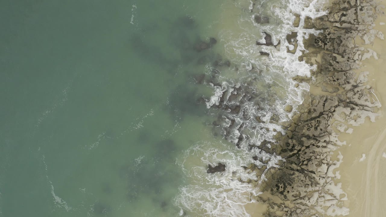 Overhead view in Baja California Sur Mexico of blue-green surf spreading over gray shoreline rocks next to tan beach sand, with no visible people.