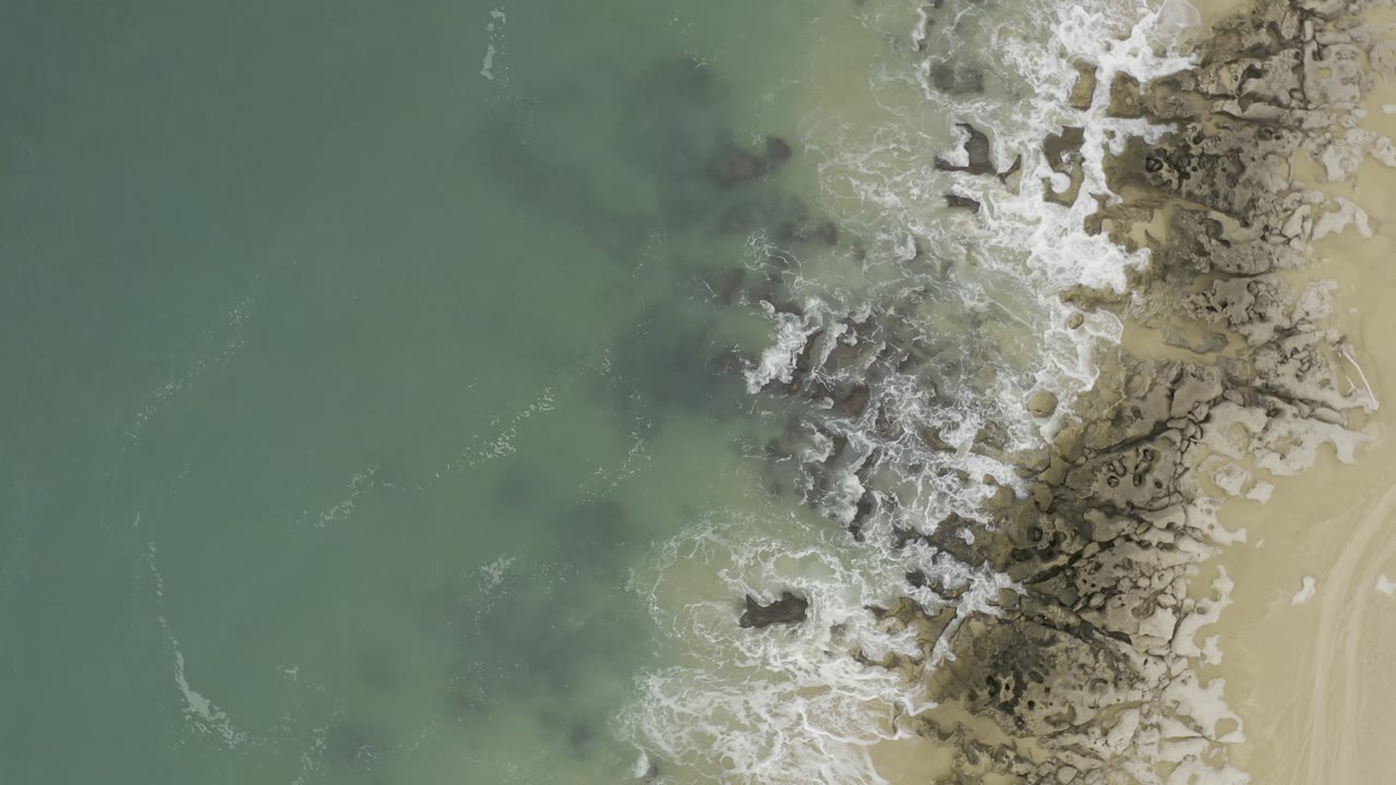 Foamy ocean water around dark rocks and sandy beach edge